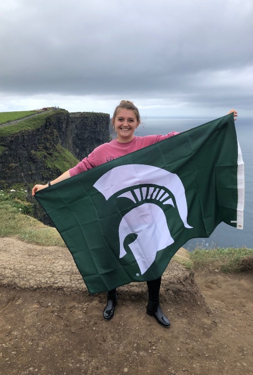 a girl on cliffs holding an MSU flag wearing a pink long sleeve, leggings, and rain boots